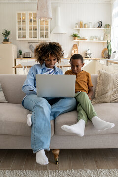 Happy African American family mother and little kid son watching cartoons on laptop together, sitting on couch at home. Sleeping mom and child looking at computer screen having video call