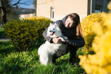 A woman is next to her white young dog in the city on a walk in spring in park, the woman is trying to train the dog but the dog enthusiastically looks away