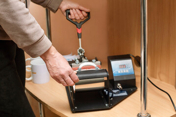 A woman makes cups to order at home, using a special thermopress to apply image printed on a sublimation printer to a cup. Production of cups with advertising images