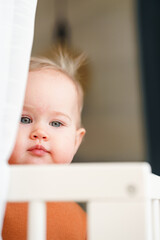 A beautiful Caucasian toddler child is standing in a crib and holding on to the white sides, top view