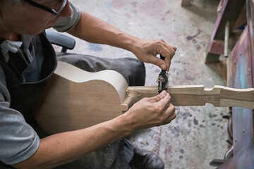middle-aged Latino man shaping the neck of a ukulele out of wood in his carpentry shop