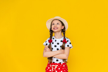 A beautiful little schoolgirl in a straw hat looks up, smiles broadly. A teenage girl in summer clothes on a yellow isolated background.
