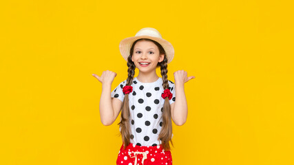 A beautiful little girl in polka dot shorts and a straw hat is pointing at your advertisement. Summer holidays. A teenage girl is waiting for summer on a yellow isolated background. Copy Space