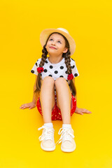 A beautiful little schoolgirl in a straw hat looks up, smiles broadly. A teenage girl is sitting in summer clothes on a yellow isolated background.