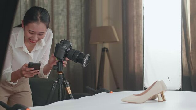 Asian Female Photographer Comparing Photos On A Smartphone To Camera While Taking Photos Of Women'S Shoes In Home Studio 
