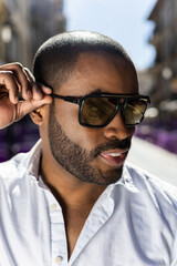 Portrait of a dark-skinned man posing with square, graduated sunglasses on the street. Promotion of...