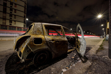 Street view of Milan with a burned down car in the middle of the street, car trails in the night...