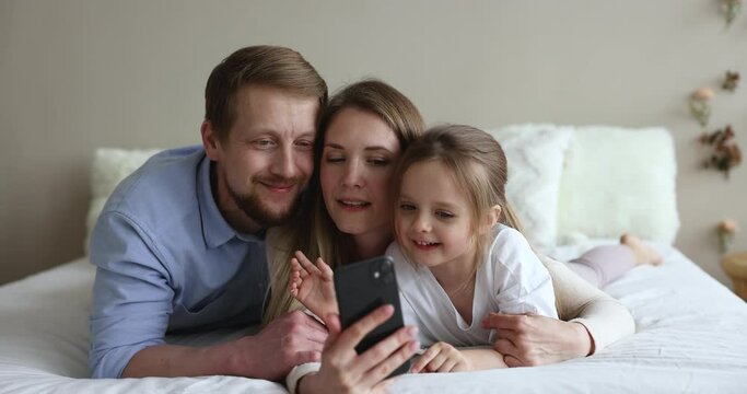 Cheerful cute daughter kid and happy couple of young parents resting on bed, taking selfie photo on smartphone, watching, shooting video for social media, making video call