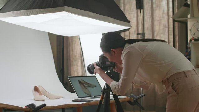 Asian Female Photographer Comparing Photos On A Laptop To Camera While Taking Photos Of Women'S Shoes In Home Studio 
