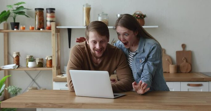 Excited millennial couple using laptop at kitchen table, chatting, receiving good surprising news, getting happy, joyful, shouting for joy, giving winner high five, hugging