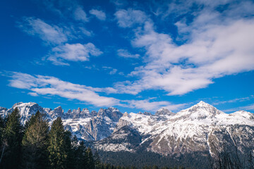 A mountain range with snow capped mountains in the background and a blue sky with clouds above it and a few trees in the foreground, Dolomiti, Italy 