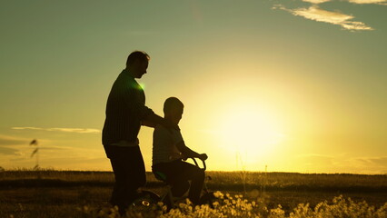 Fototapeta na wymiar Active family, dad teaches his son to ride bike in park at sunset. Baby daddy play outside. Father teaches little boy to ride childrens bicycle on road in countryside in sun. Happy family, childhood