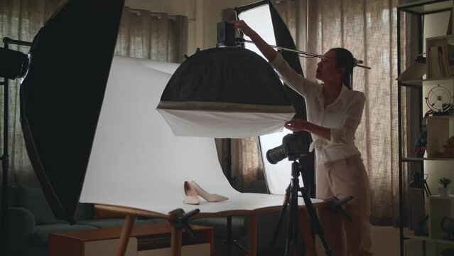 Asian Female Photographer Adjusting Light Equipment To Get Good Illumination On Women'S Shoes While Taking Photos Of The Products In Home Studio 
