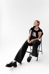 Fototapeta premium Full body of young female with leather jacket on shoulders and blond hair sitting on chair and relaxing against white background