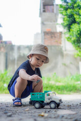 little child playing with toy car on the playground. asian kid boy play outside in backyard on sunny morning wearing brown hat and black shirt