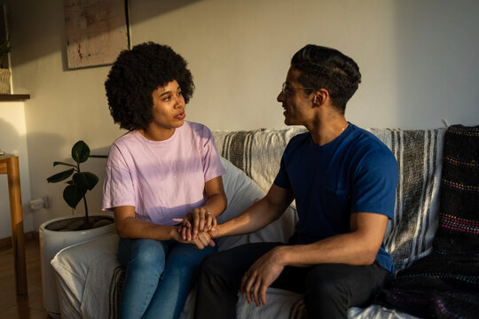 Two Latin Friends Having A Serious Conversation. They Are Holding Hands And Siting On The Sofa. 