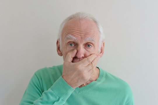 Close-up portrait of an elderly gray-haired man with an expressive, stunned look and his mouth covered in surprise with his hands on a light background,