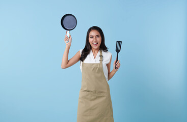 Happy asian woman chef holding spatula and frying pan utensils cooking in the kitchen on blue...