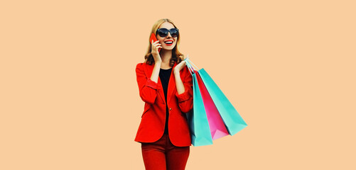 Portrait of beautiful smiling young woman calling on smartphone with shopping bags wearing business blazer on pink background