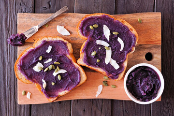 Toast with ube halaya jam, coconut and pumpkin seeds. Top down view platter on a dark wood background. Sweet, trendy, purple yam spread.