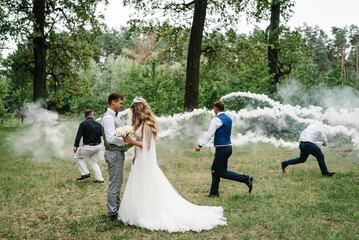 Newlyweds and their friends raise their hands up posing on the park. Running with smoke bombs.