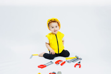A little boy 2 years old in a builder's suit on a white background. A child in a plastic helmet and plastic tools. Plastic children's toys.