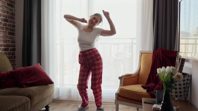 Happy smiling woman in pajamas and slippers dancing against big window in living room. People relaxing at home, having fun, positive emotions and joy