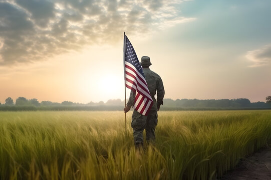 Military Soldier in miitary gear from behind in a sunny wheat field landscape holding an american flag