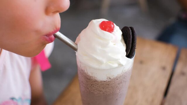 Close-up of little girl drinking chocolate milkshake or cocktail with berry and cookie on top of it at the cafe or restaurant, slow motion