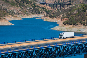 Truck with a refrigerated trailer driving on a metal bridge over a blue water swamp.