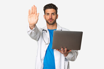 Young doctor man holding a laptop standing with outstretched hand showing stop sign, preventing you.