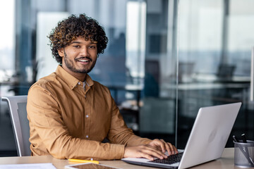Portrait of young successful Indian man at workplace inside office, businessman smiling and looking...