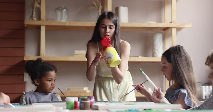 Diverse group of artistic school kids painting on class. Cute boy stroking acrylic, oil colors on canvas, speaking in studio, training skills. Art teacher helping children to add paint from bottle