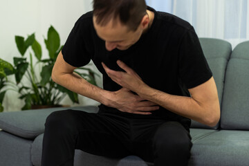 Man suffering with severe stomach pain sitting at home. Hand of mature guy holding abdomen suffering from ache, diarrhea or indigestive problem. Caucasian guy pressing on belly on painful sensation