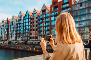 Back view of young European lady tourist in hat makes photo or video on smartphone at city of Gdansk, Poland, Renovated granaries on Granary Island on river Motlawa in Gdansk, Poland