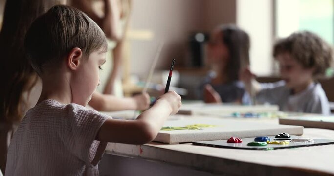 Focused cute boy practicing skills on drawing class in artistic school, sitting at table, painting with paintbrush on canvas at palette, studying with group in creative studio, children club