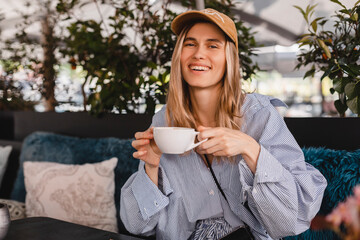 Beautiful young woman looking at camera with coffee sitting in cafe and look happy. Blonde wearing stripped shirt and beige cap sitting on the sofa in restaurant. Coffee break.