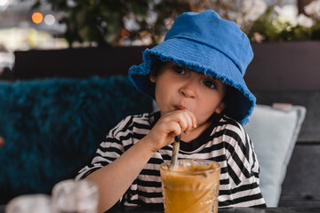 A boy sits in a cafe and drinks apple juice from a tubule. Boy wear blue panama hat and stripped black and white t-shirt.