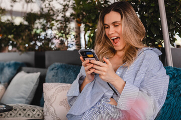 Happy smiling blonde woman looking at smartphone in her hand with excited face expression, unable...