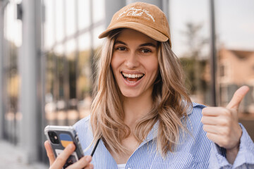 Beautiful smiling fashionable woman sitting standing in front of business center and show thumb up, hold mobile phone, look happy. Amazed woman wear beige cap, white and blue shirt.
