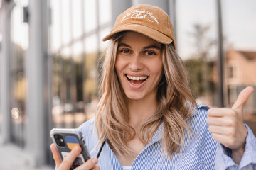 Beautiful smiling fashionable woman sitting standing in front of business center and show thumb up, hold mobile phone, look happy. Amazed woman wear beige cap, white and blue shirt.