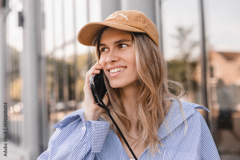 Wall mural Closeup portrait of a cheerful young woman making a phone call outdoors in the city walking near office. Happy woman wear stripped shirt and beige cap. Female look at side.