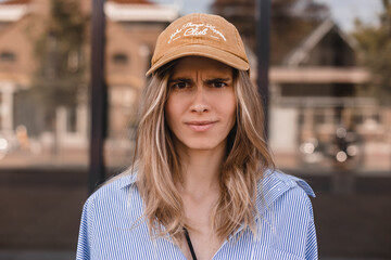 Drama blonde curly woman look angry at camera walking on the street. Girl wear beige cap and stripped shirt. Portrait of a wtf angry young girl wearing casual clothing standing near office building.