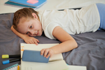 Smiling girl lying on bed and watching entertaining videos on smartphone insted of doing homework