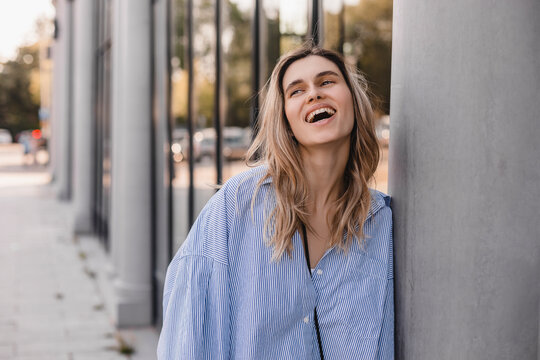 Cute young woman with a lovely sense of humor standing leaning against a glass window exterior column with copy space in an urban street laughing at the camera. Attractive blonde girl wear shirt.