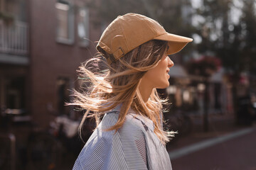 Close up photo of young blonde woman walking outdoor on the street in city, wear cap, stripped shirt, flying hair. Woman look happy and smiling.