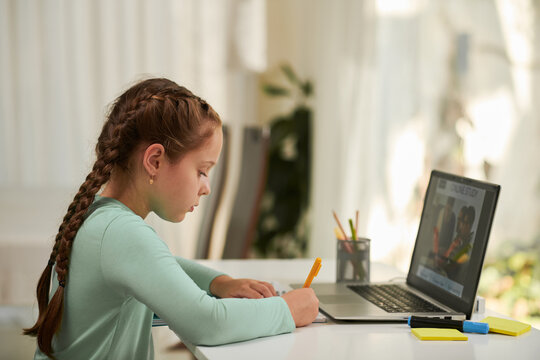 Schoolgirl taking notes in textbook when watching educational video on school platform