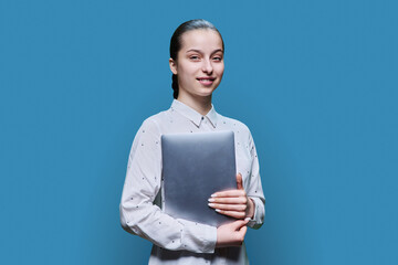 Teenage girl holding closed laptop in her hands, on blue studio background