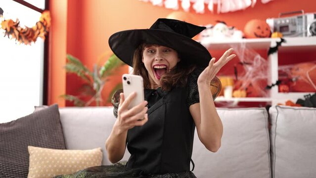 Young caucasian woman wearing witch costume sitting on the sofa with smartphone at home