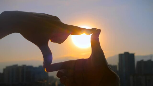 Silhouette of woman hands are doing finger frame and looking at the sunset, sunrise sky over the city - close up view, sun lens flares. Gesture, imagination, creativity and composition concept